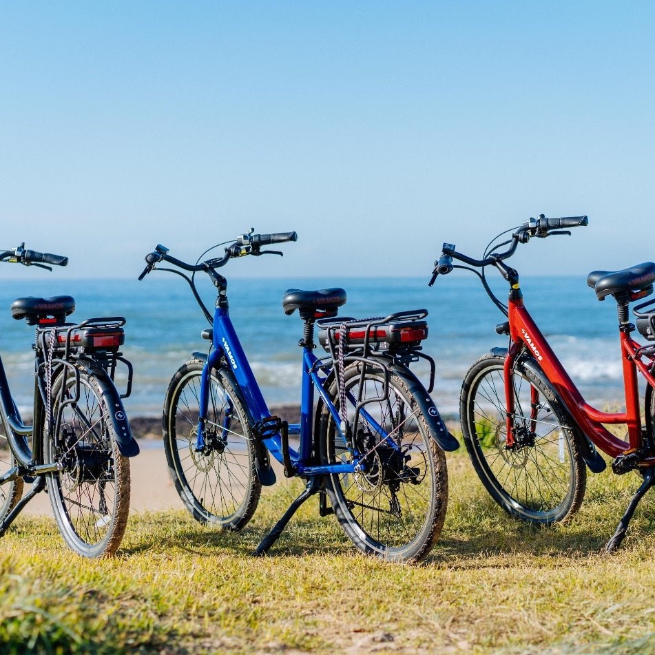 Vamos El Rapido step-through electric bicycle featuring a bright blue frame parked next to a red model on a grassy coastal path.