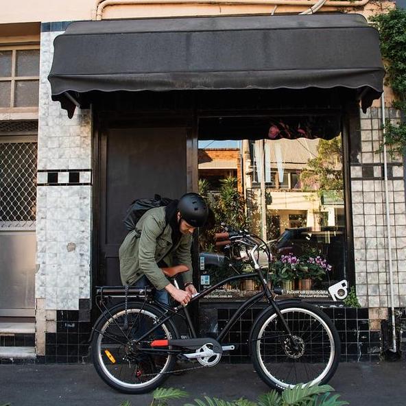 A person adjusting a black cruiser-style electric bicycle with a glossy frame, swept-back handlebars, and a rear rack